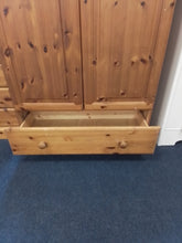 Load image into Gallery viewer, A light natural pine wooden storage cabinet with two upper doors and one open drawer below, set against a blue carpeted floor.